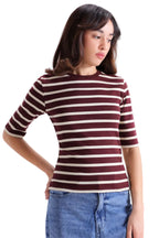 A woman wearing a short-sleeve crewneck t-shirt with horizontal burgundy and cream stripes, paired with blue jeans, photographed against a light gray studio background.