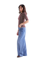 A woman modeling a short-sleeve crewneck top with dark red and white horizontal stripes, paired with wide-leg light blue jeans, shown from a side profile against a light gray studio background.