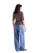 A woman shown from the back wearing a brown and white striped short-sleeve top and wide-leg blue jeans, standing against a light gray studio background.